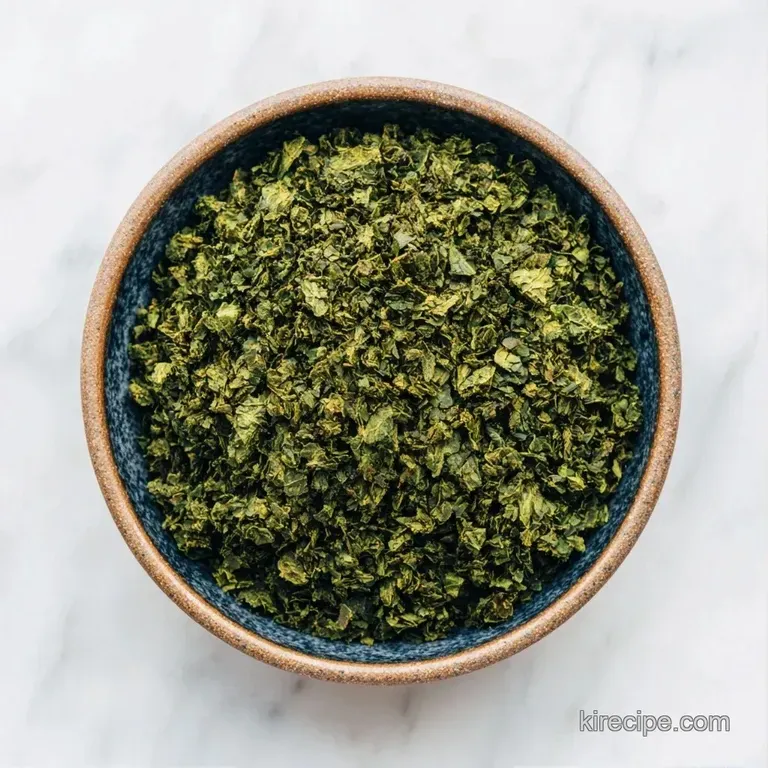A small crystal bowl of forest-green dried herbs beside a fresh basil sprig on a clean, white marble surface.